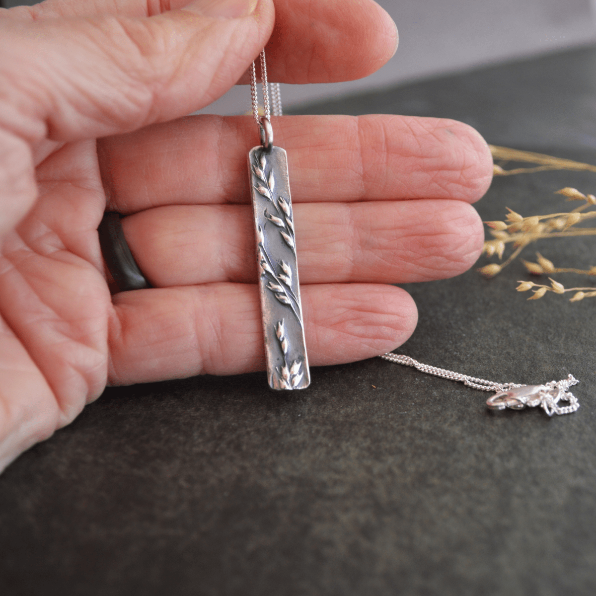 A Hand Holding A Long Silver Pendant Textured with Prairie Switchgrass - Gayle Dowell