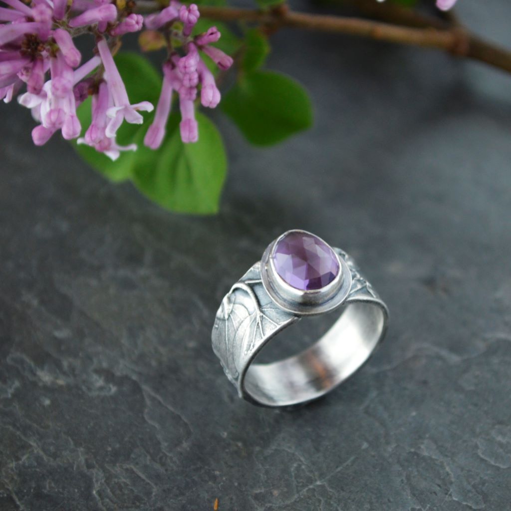 Sterling silver ring with a texture of a lilac plant.  Bezel-set purple amethyst tear-drop shaped gemstone sits on top.  Lilac flowers and leaves in the backround behind the ring.  